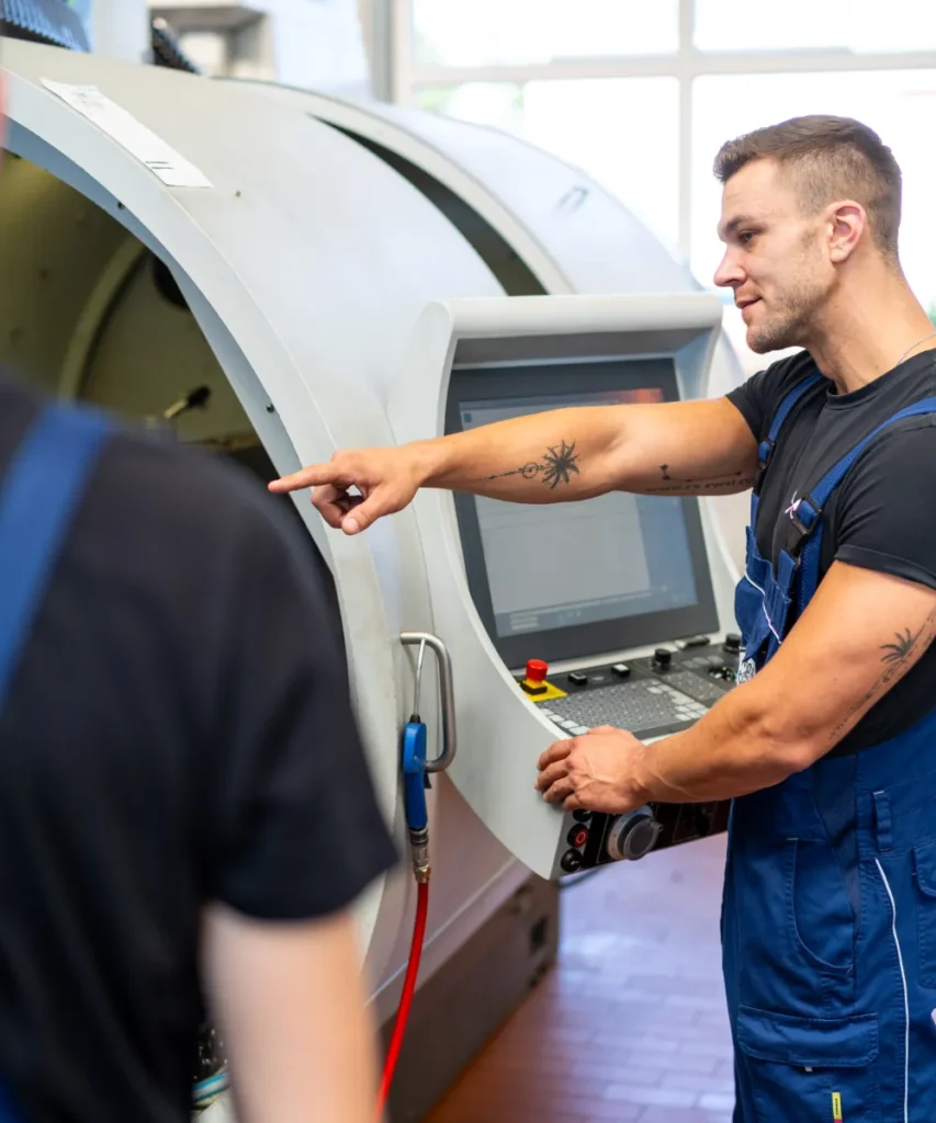 Mitarbeiter erklärt einem Kollegen die Bedienung einer CNC-Drehmaschine am Steuerungspanel